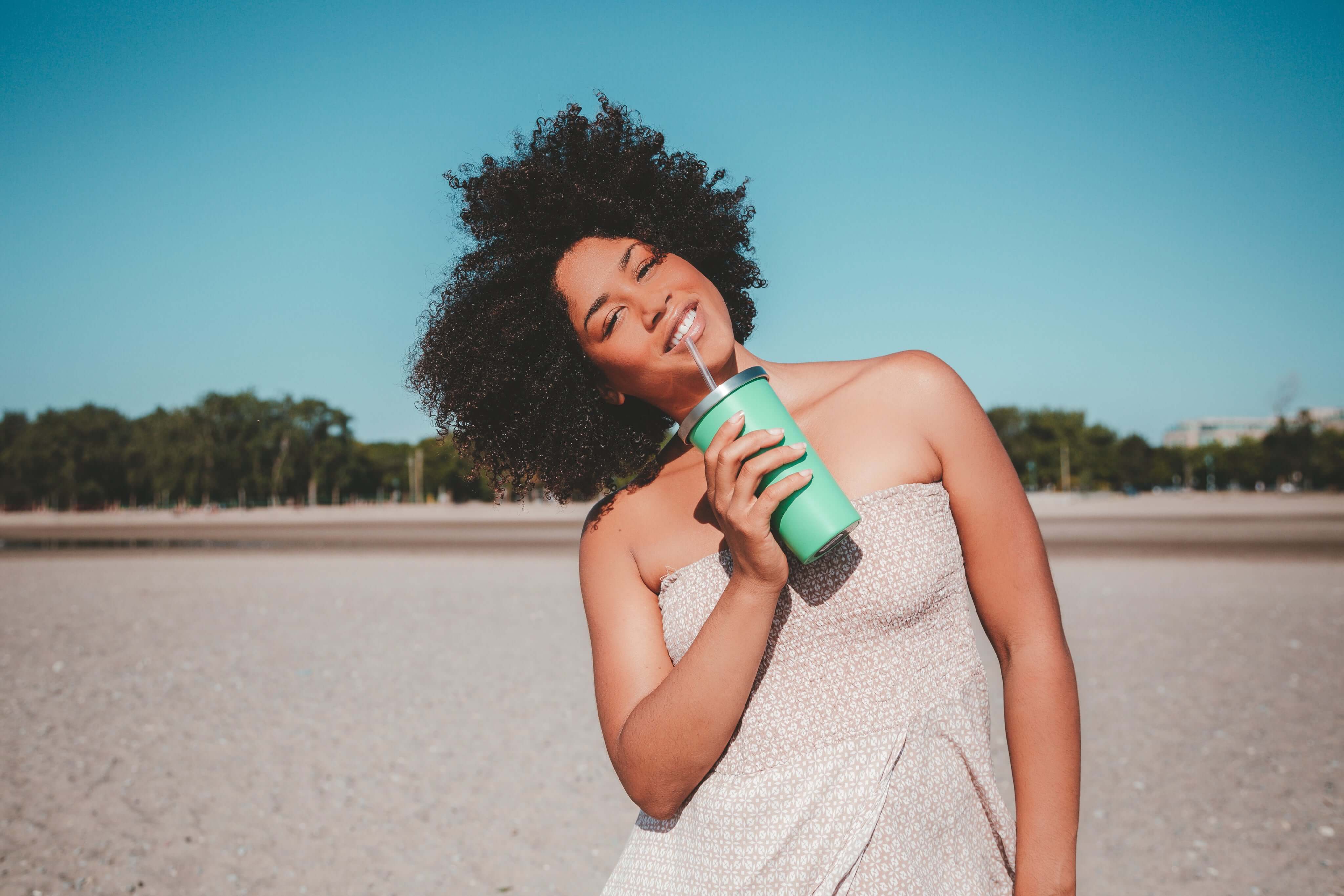 Woman enjoying a refreshing drink at the beach, smiling in a sunny outdoor setting with a carefree vibe.